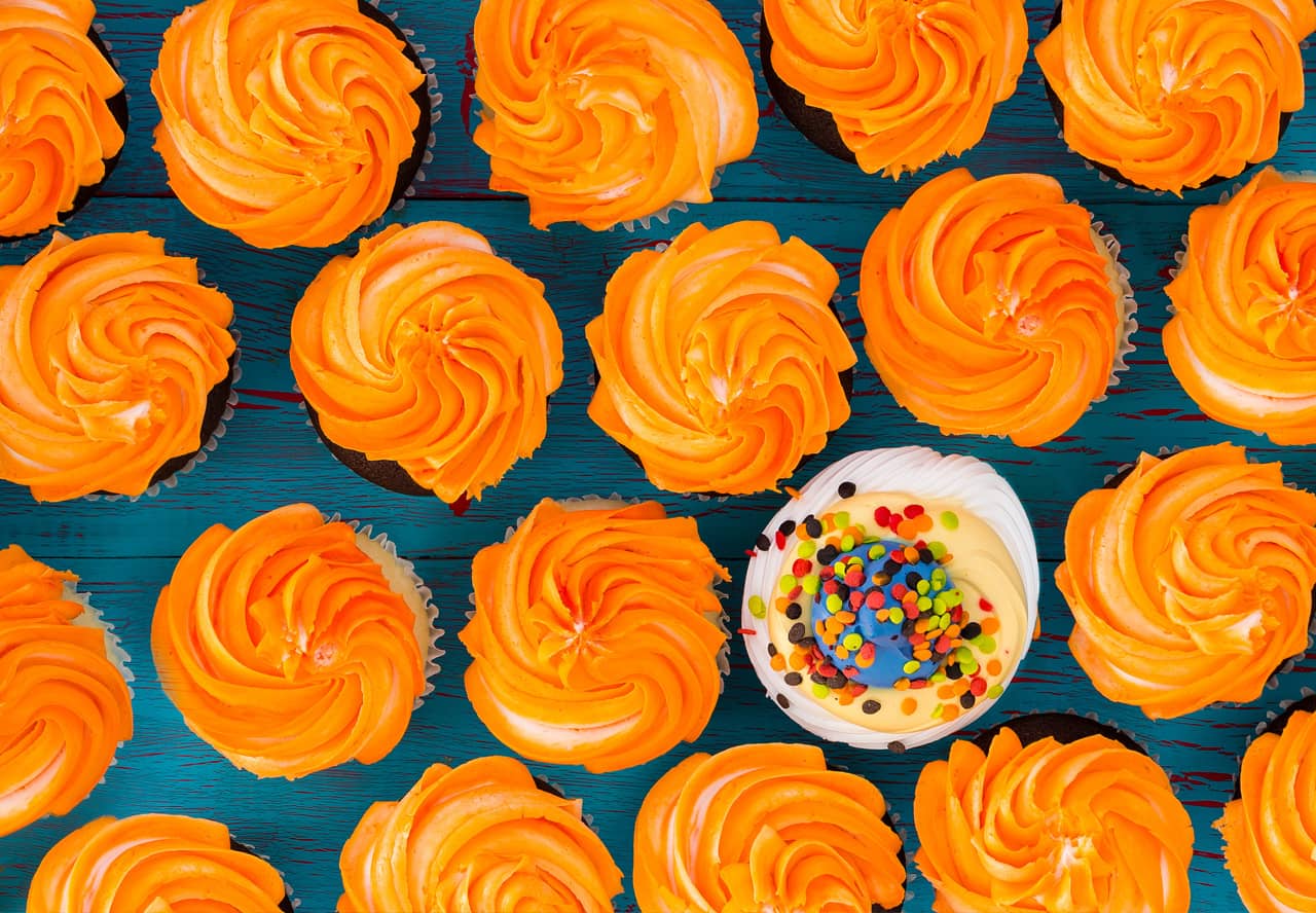A photo of delicious cupcakes with orange frosting, but one cupcake has white and light orange frosting and sprinkles representing the concept of being different