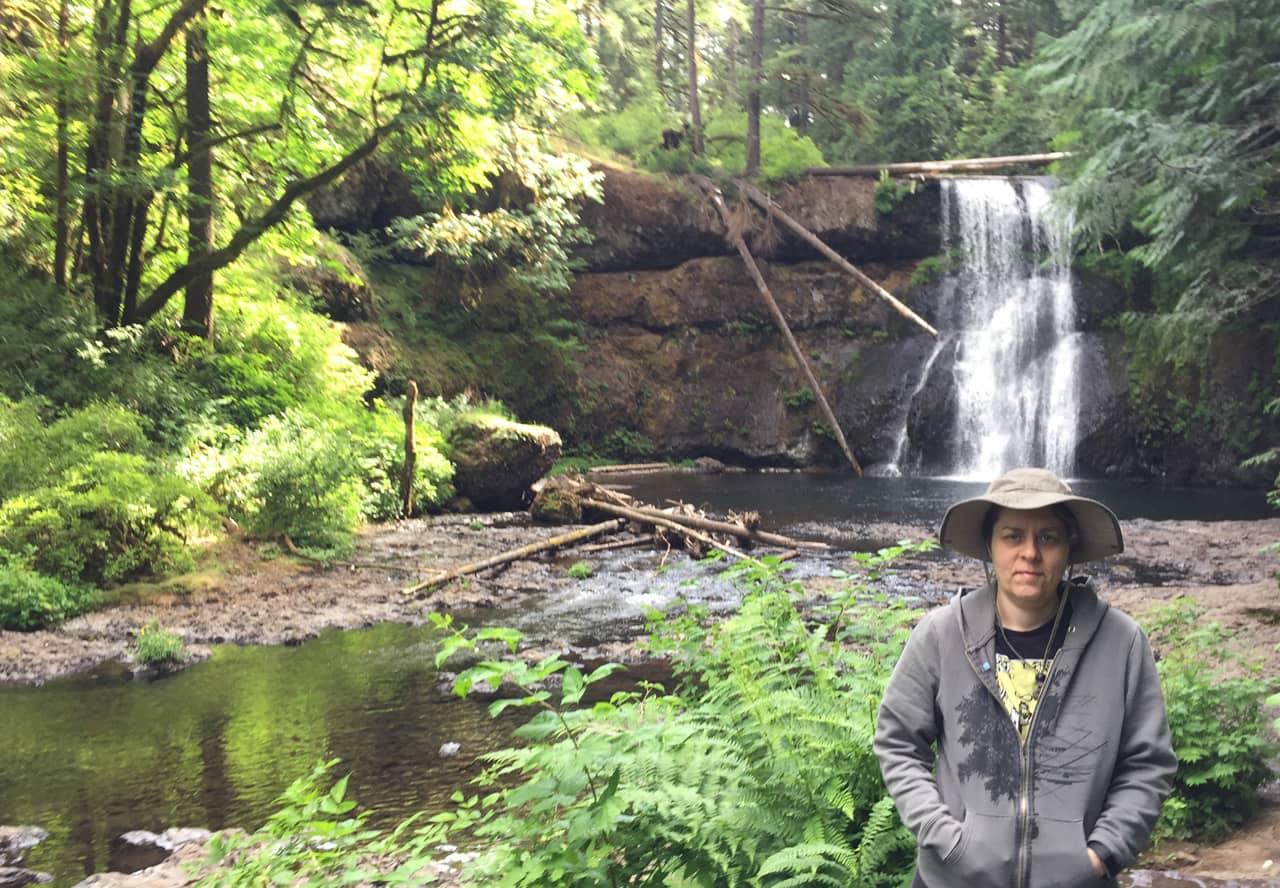 A photo of Sherry Holub in front of a waterfall surrounded by green trees and foliage