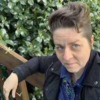 A photo of Sherry Holub leaning on the railing of some wooden steps in front of a large holly bush